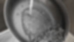 Water pouring into a black frying pan, creating ripples and bubbles. The pan glistens with droplets, set in a kitchen sink.